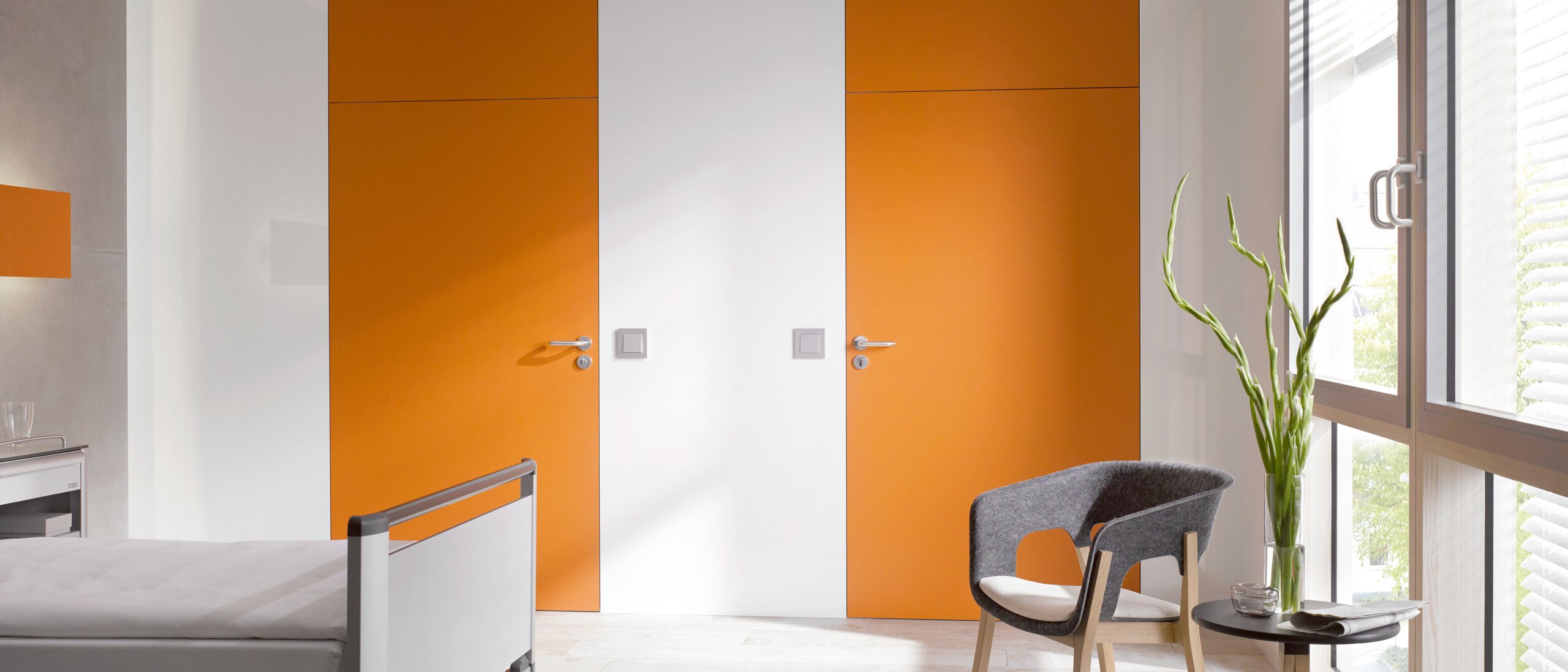Weißes Krankenhauszimmer mit 2 orangenen Türen ausgestattet mit HEWI Türdrücker 111 in Grau aus Polyamid