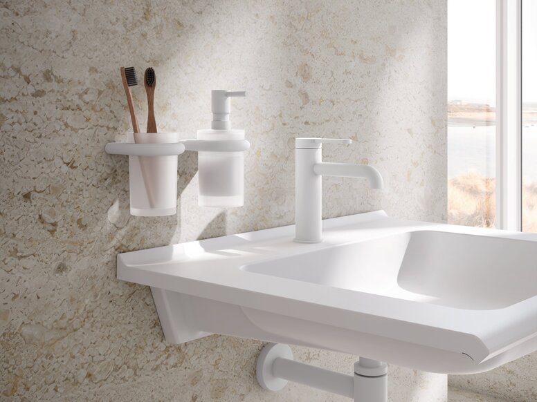 HEWI Armaturen AQ477 Weißer Wandbecher mit Zahnbürsten und weißem Seifenspender über einem Waschbecken mit Wasserhahn vor beigefarbener Steinwand