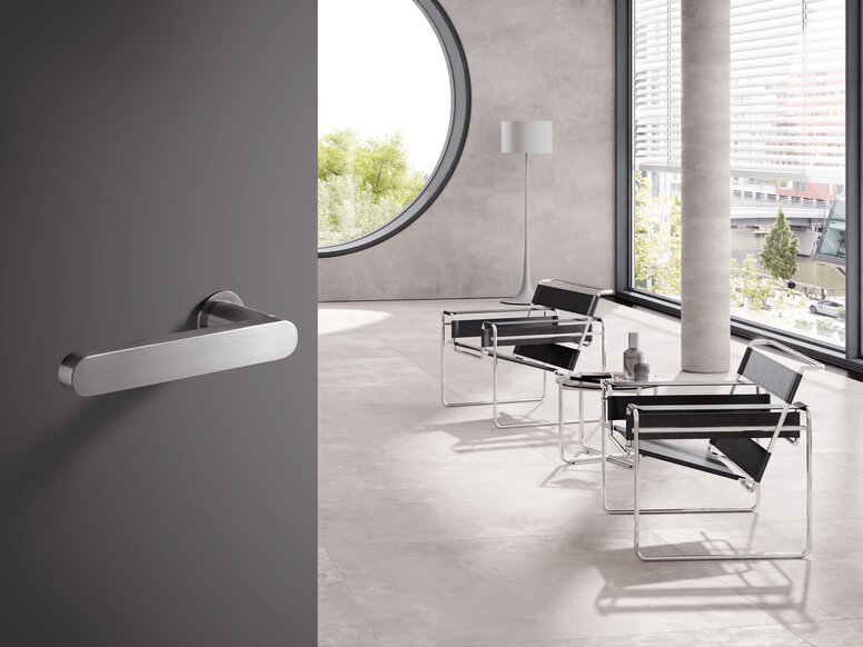 HEWI Serie 270 Türdrücker Türklinke aus Edelstahl an grauer Tür, dahinter heller Raum mit Stühlen