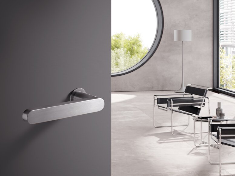 A stainless steel door handle on a grey door; behind it, a bright room with chairs