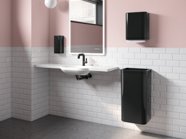 HEWI Range 477 Interior A white washbasin with a black paper towel dispenser and wastebin next to it, in a bathroom with white tiles and pink walls