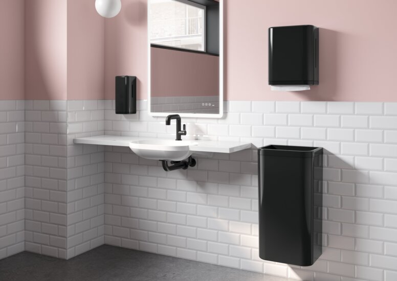 HEWI Range 477 Interior A white washbasin with a black paper towel dispenser and wastebin next to it, in a bathroom with white tiles and pink walls