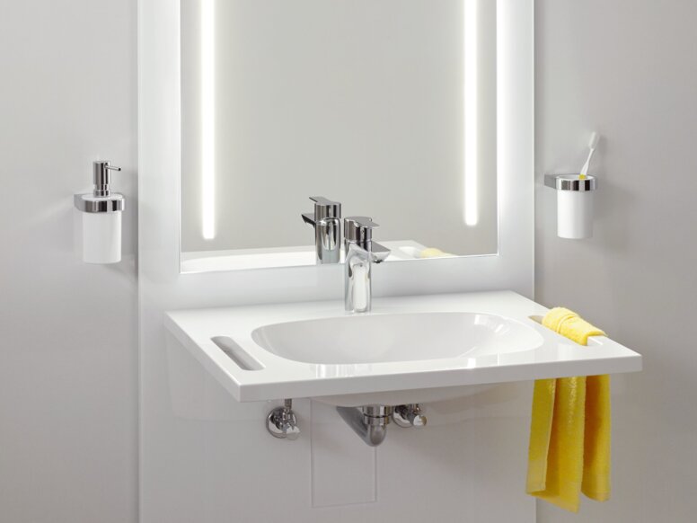 HEWI S 50 Washbasin Unit A white washbasin with a tap, a recessed handle on the side and an illuminated mirror with two light strips on a light grey wall