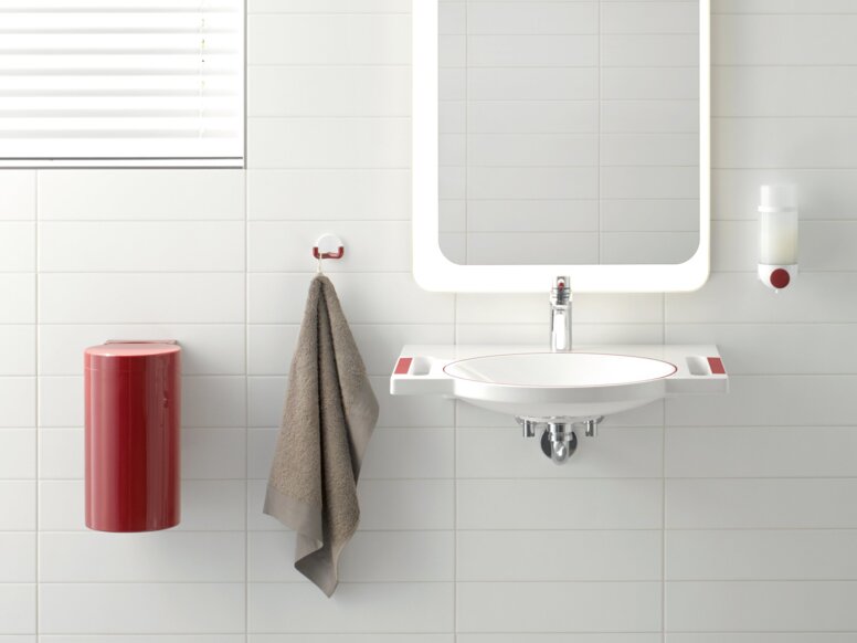 A red hinged support rail next to the WC with a red cover, alongside a white washbasin with red recessed handles, and an illuminated mirror above it