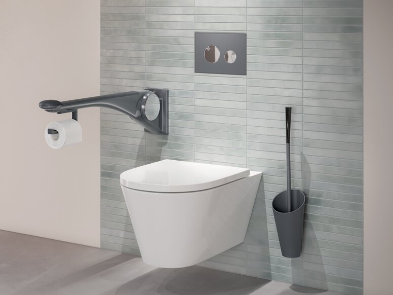 HEWI Range 802 LifeSystem premium hinged support rail handle Wall-hung WC with a side support rail, an integrated toilet roll holder and a toilet brush mounted on a green tiled wall