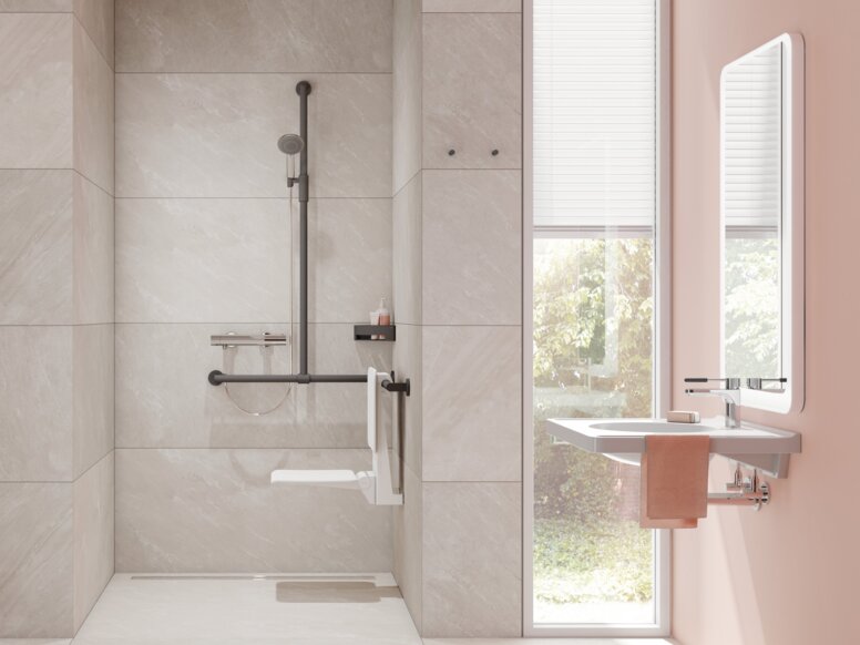 A hinged seat with a backrest and a rail with shower head holder in an accessible bathroom with a wheelchair-accessible washbasin in front of a window wall