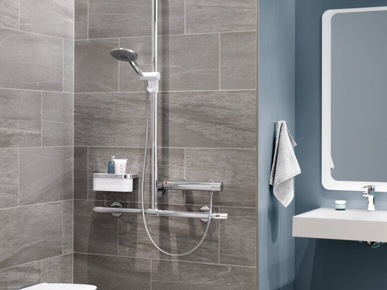 A hinged shower seat with a support rail, a thermostatic mixer and a shower head against a grey tiled wall, next to which is a washbasin area against a blue wall