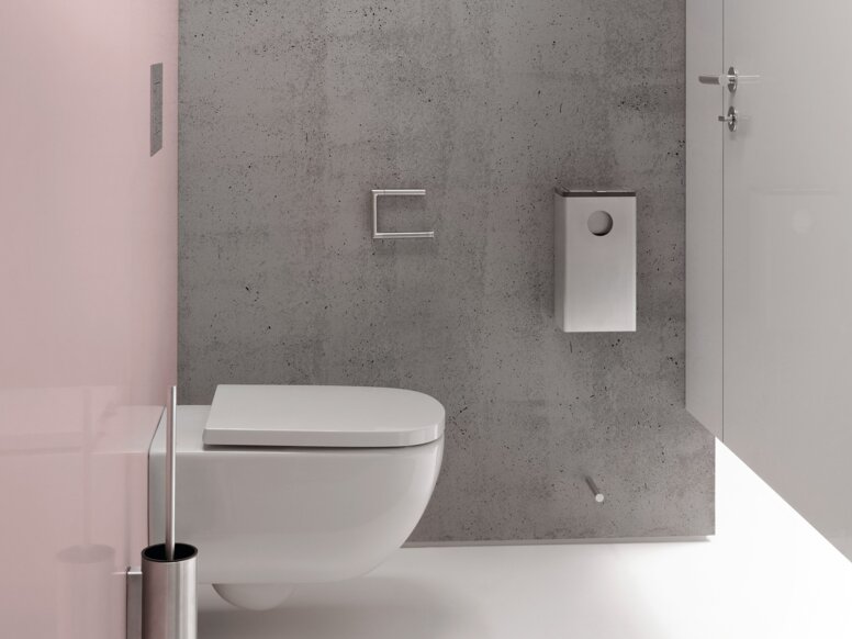 HEWI System 162 Interior Narrow toilet area with a white wall-hung toilet against a grey concrete-effect wall, a white toilet roll holder and toilet brush set, and a pink side wall