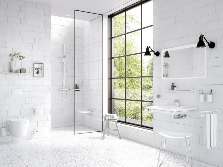 HEWI System 162 Interieur Helles Badezimmer mit weißen Wandfliesen, bodengleicher Dusche hinter einer Glasabtrennung, wandhängendem WC und weißem Waschtisch unter einem Spiegel, ergänzt durch weiße Haltegriffe und ein großes schwarzes Sprossenfenster