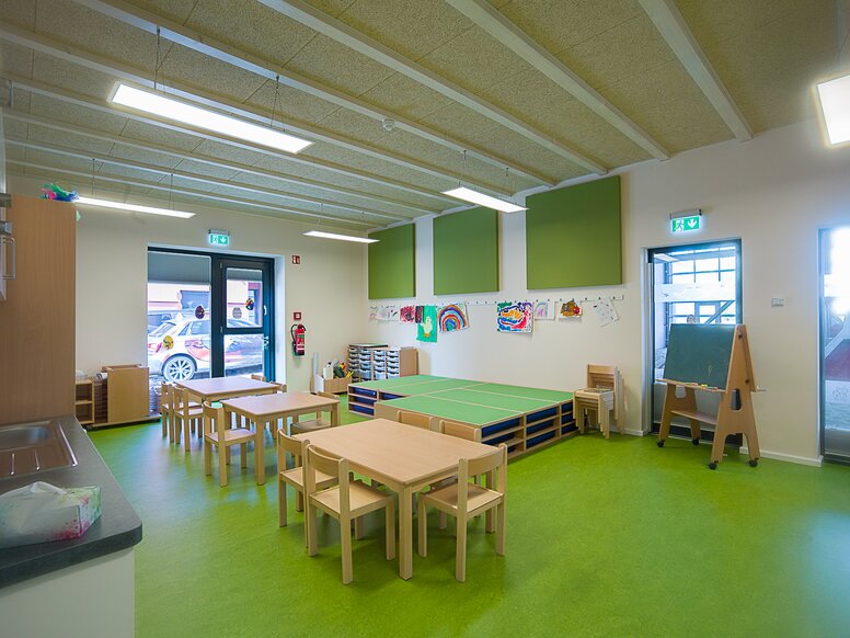 Gruppenraum einer Kindertagesstätte mit grünem Boden und Tischen und Stühlen aus Holz